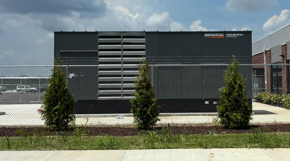 Generac Industrial Power generator unit installed behind a chain-link fence with small evergreen trees in front