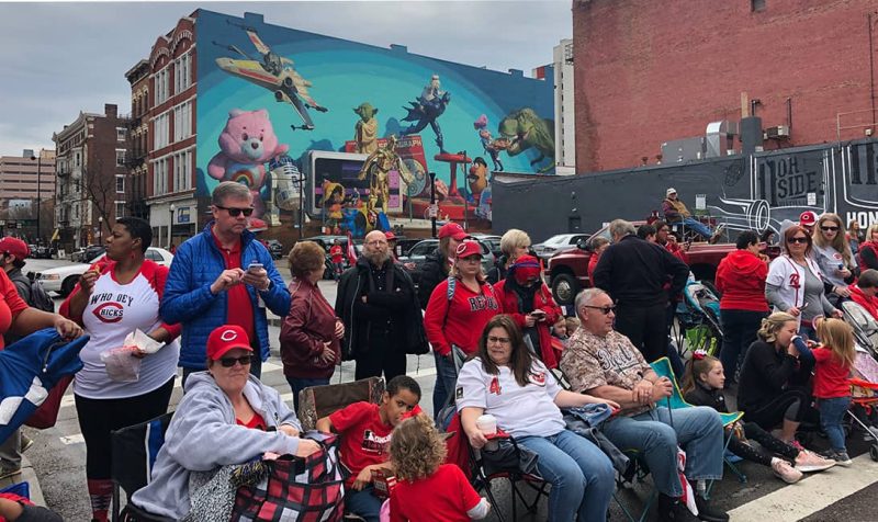 Spectators in Cincinnati gather on a city street in front of a large pop culture mural